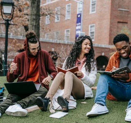 Full body of happy diverse students with notebooks and laptop sitting on grassy lawn on campus of university while studying together