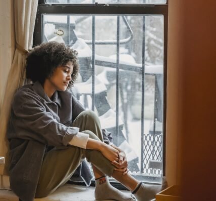 Side view full body of upset African American woman embracing knees while sitting in solitude near window and looking down